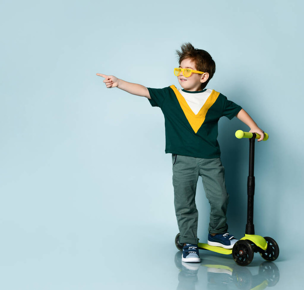 Boy in colorful t-shirt, yellow sunglasses, jeans, sneakers. Screaming with raised hand, standing on kick scooter. Blue background - Foto, immagini