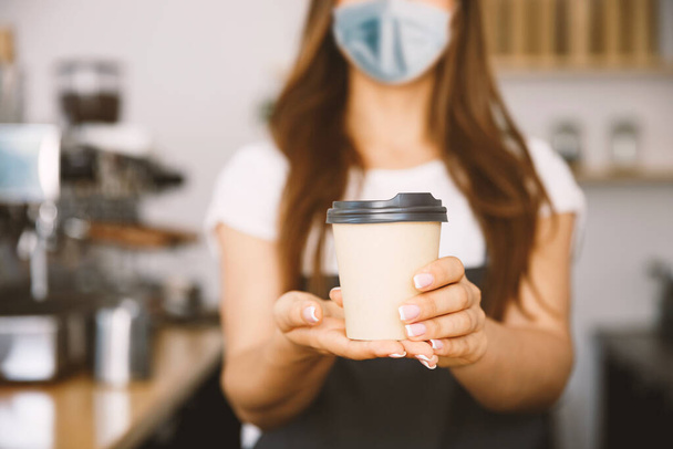 Business Owner Concept - Schöne kaukasische Barista in Gesichtsmaske bietet Einweg-Kaffee zum Mitnehmen im modernen Coffeeshop - Foto, Bild
