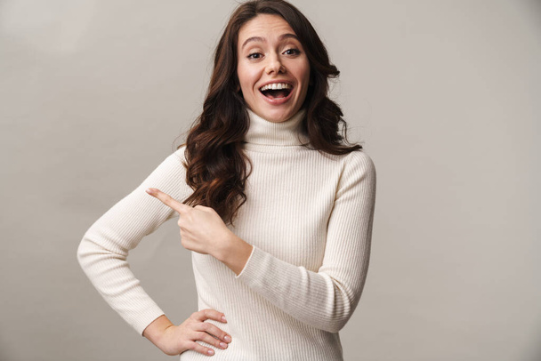 Photo of caucasian beautiful woman with long brown hair smiling and pointing finger aside at empty space isolated over beige background - Foto, immagini