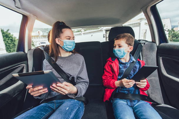 Kids wearing anti virus masks and using digital tablets in the car. Kids are travelling in car during coronavirus outbreak - Photo, Image