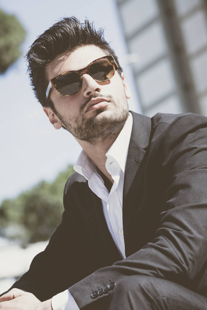 Уверенный молодой успешный человек, сидящий в городе. В солнечных очках, позади него современное офисное здание. Модные волосы, белая рубашка и куртка
. - Фото, изображение