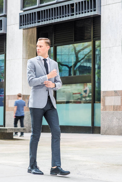 Mladý evropský podnikatel cestující po New Yorku, v šedém saku, bílé košili, kravatě, černých kalhotách, kožených botách, rukou se dotýkajících manžetách, procházející se po ulici před kancelářskou budovou, rozhlížející se - Fotografie, Obrázek