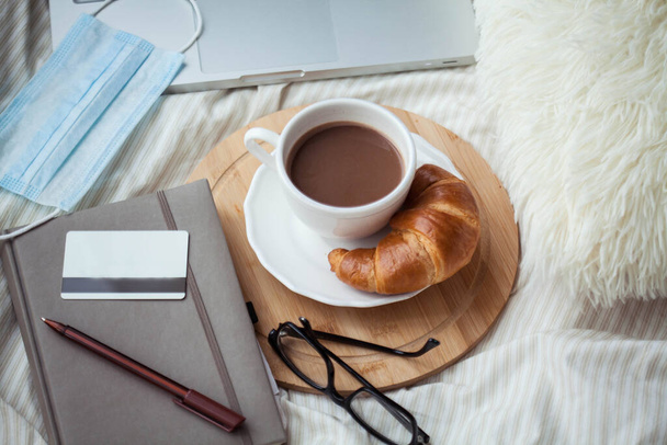 A cup of hot cocoa, a croissant, a wooden stand, a medical mask, a notebook, a laptop, glasses, a bank card on the bed. Quarantine time. Home Office. - Photo, Image