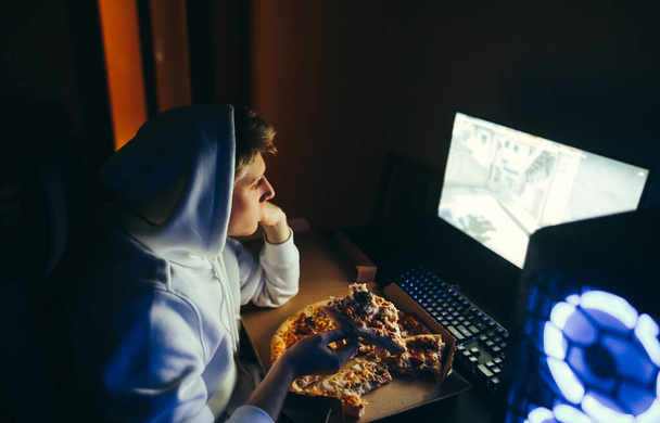 Young man sits at night at a table, watches a video on a computer and eats pizza from a hoarder. Guy teenager in a hoodie booze having pizza at the computer. Fast food and gamer. - Photo, Image