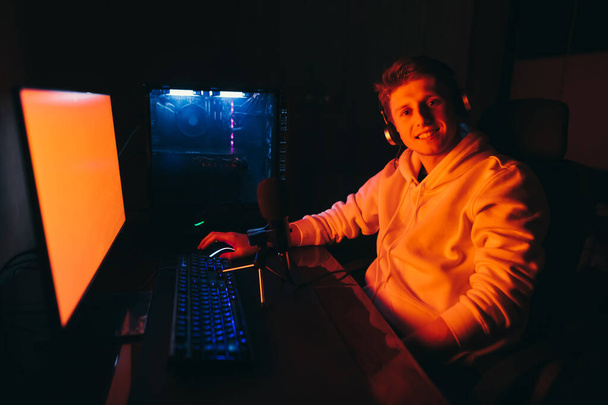 顔に笑顔で、カメラを見てコンピュータの近くの机に座っている赤い光の中で幸せなゲーマーの肖像画。陽気な若い男はコンピュータゲームをプレイし、カメラにポーズ. - 写真・画像