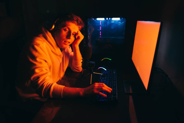 眠い若い男がコンピュータで夜に座って、悲しい顔をしたカメラを見ている。自宅でテーブルで疲れたゲーマーの肖像画、カメラを見て、眠りに落ちる。眠いゲーマーはゲームをプレイ - 写真・画像