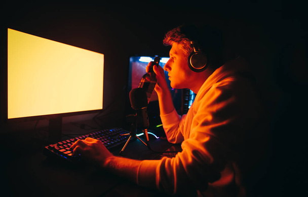 Портрет молодого человека в наушниках и с микрофоном, играющего в видеоигры дома ночью, смотрит на красный экран и держит в руке пивную бутылку. Геймер играет в компьютерные игры и пьет пиво
 - Фото, изображение