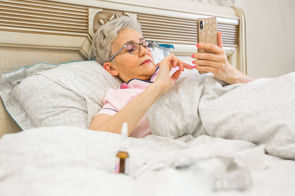 an elderly woman lies on a bed with a phone - Fotoğraf, Görsel