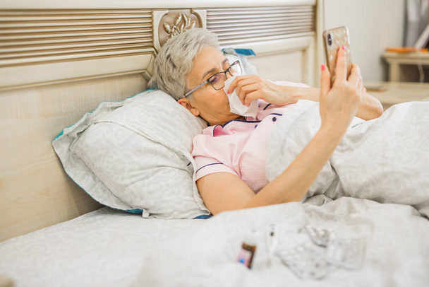 Yaşlı bir kadın telefonu olan bir yatakta yatıyor. - Fotoğraf, Görsel