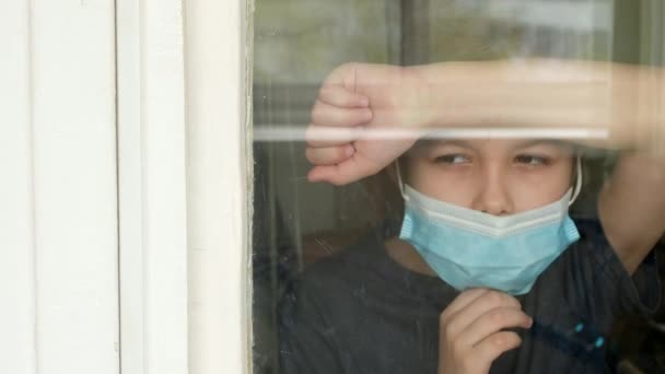 Ένα νεαρό αγόρι με ιατρική μάσκα κοιτάει έξω από το παράθυρο. Αυτοαπομόνωση σε καραντίνα, coronavirus, covid 19. - Πλάνα, βίντεο