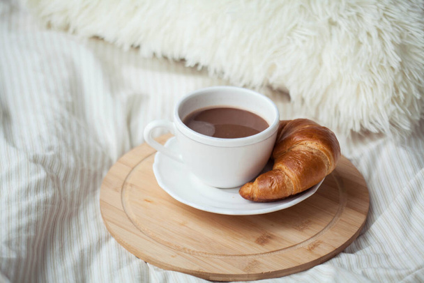 Egy csésze forró kakaó, egy croissant, egy faállvány az ágyon. Karantén idő. A hajléktalanság. Hangulatos. - Fotó, kép