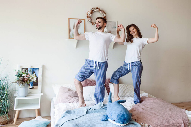 Ein junges Paar überprüft seinen aufgeblasenen Bizeps, während es im Schlafanzug auf dem Bett steht - Foto, Bild