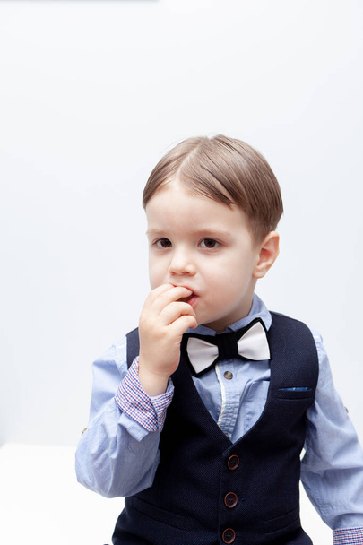 A boy in a shirt, vest and trousers is eating a delicious candy. White background. The child is smart. Hair parted in the middle - Foto, Imagen