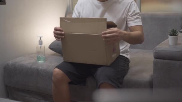 Asian male's unboxing cardboard box, on seven sofa home, mail receiver unpack small carton parcel packel packel package, covid-19 Crisis, доставка пакетів онлайн купівлі, розпакувальний пакет, відправка пошти - Кадри, відео