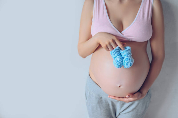 Изолированный портрет неузнаваемой беременной женщины с голубыми вязаными кроссовками и с большим животом. Беременность, материнство, нерожденность и концепция ожидания. Свободное место для макета текста
 - Фото, изображение