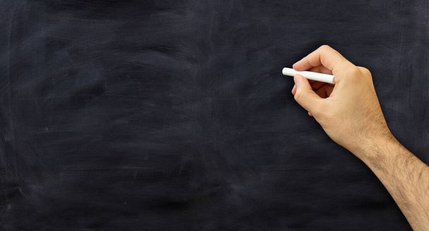 Man hand with a chalk isolated on black background. Teacher holding a white chalk against a chalkboard, copy space - Photo, Image