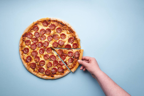 Woman's hand grabbing a slice of pepperoni pizza. Delicious homemade pizza with an abundance of meat. Sliced pizza salami, above view - Photo, Image
