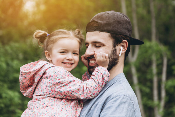 stylish bearded father with his little daughter in his arms against the backdrop of green trees on a walk in the park hugging and kissing a baby - Foto, immagini
