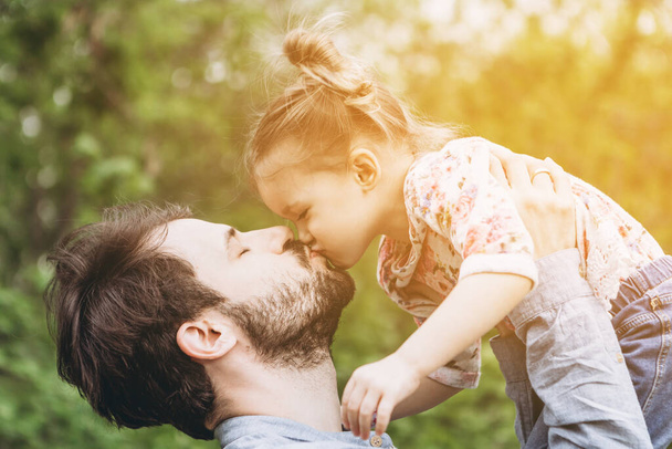 stylish bearded father with his little daughter in his arms against the backdrop of green trees on a walk in the park hugging and kissing a baby - Foto, Bild