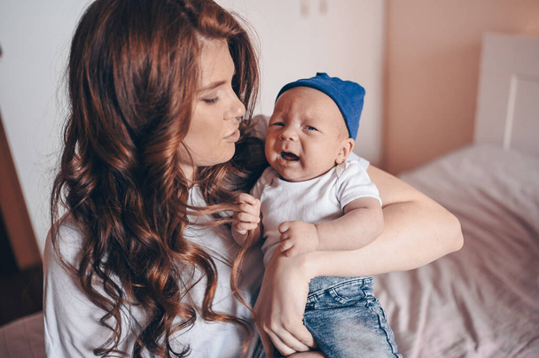 Close up portrait of happy young mom holding her crying baby in blue jeans and white t-shirt and cap. Young happy family, mother playing with cute emotional little newborn child son in the bedroom. - Zdjęcie, obraz