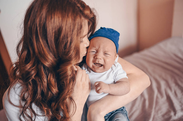 Primer plano retrato de la feliz mamá joven sosteniendo a su bebé llorando en vaqueros azules y camiseta blanca y gorra. Joven familia feliz, madre jugando con el pequeño hijo recién nacido emocional lindo en el dormitorio
. - Foto, imagen