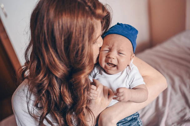 Primer plano retrato de la feliz mamá joven sosteniendo a su bebé llorando en vaqueros azules y camiseta blanca y gorra. Joven familia feliz, madre jugando con el pequeño hijo recién nacido emocional lindo en el dormitorio
. - Foto, imagen