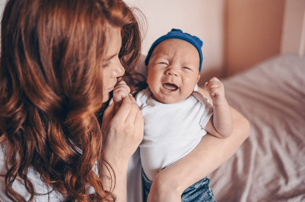 Close up portrait of happy young mom holding her crying baby in blue jeans and white t-shirt and cap. Young happy family, mother playing with cute emotional little newborn child son in the bedroom. - Foto, imagen