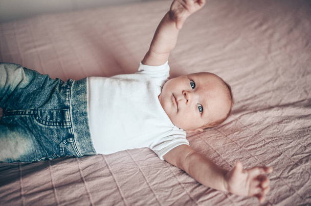 Aranyos kisfiú, rózsaszín takarón fekszik a hálószobában. Újszülött. A baba ébren marad az ágyon. Szoros portré az érzelmes újszülött babáról kék farmerban, kék sapkában és fehér pólóban - Fotó, kép