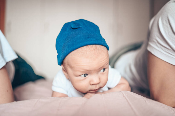 Милый маленький мальчик лежит на розовом одеяле в спальне. Новорожденный. Ребенок не спит на кровати. Крупный план портрета эмоционального новорожденного ребенка в синих джинсах, голубой кепке и белой футболке
 - Фото, изображение