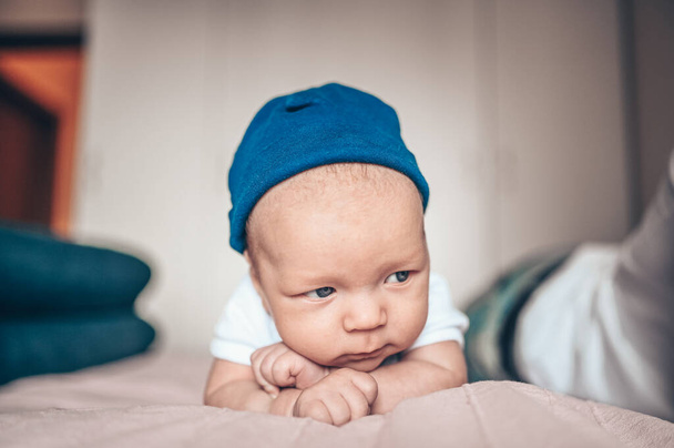 Cute little baby boy lying on pink blanket at bedroom. Newborn. Baby stays awake on the bed. Closeup portrait of emotional newborn baby in blue jeans, blue cap, and white t-shirt - Foto, imagen