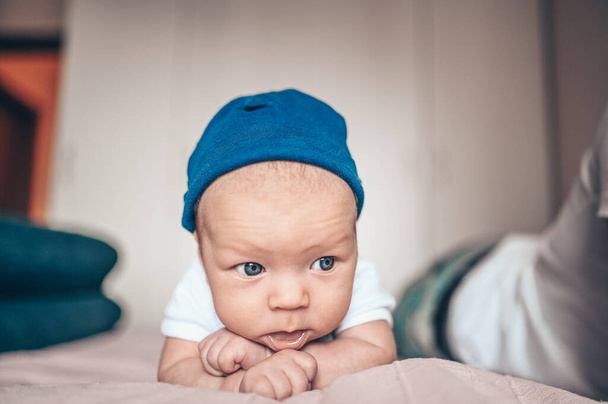 Cute little baby boy lying on pink blanket at bedroom. Newborn. Baby stays awake on the bed. Closeup portrait of emotional newborn baby in blue jeans, blue cap, and white t-shirt - Фото, изображение