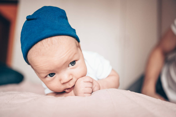 Милый маленький мальчик лежит на розовом одеяле в спальне. Новорожденный. Ребенок не спит на кровати. Крупный план портрета эмоционального новорожденного ребенка в синих джинсах, голубой кепке и белой футболке
 - Фото, изображение