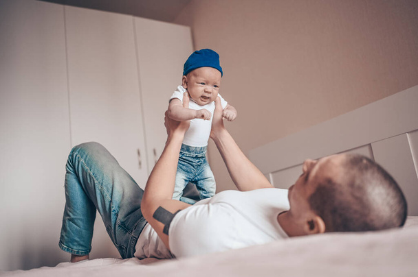 Close up portrait of happy young dad father holding his baby in blue jeans and white t-shirt and cap. Young happy family, dad playing with cute emotional little newborn child son in the bedroom. - 写真・画像