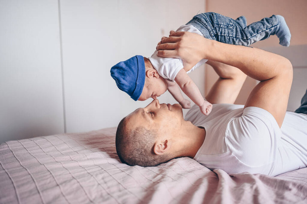 Κοντινό πορτραίτο του νεαρού πατέρα που κρατάει το μωρό του με μπλε τζιν και λευκό μπλουζάκι και καπέλο. Νεαρή ευτυχισμένη οικογένεια, μπαμπάς παίζει με χαριτωμένο συναισθηματική λίγο νεογέννητο παιδί γιο στην κρεβατοκάμαρα. - Φωτογραφία, εικόνα
