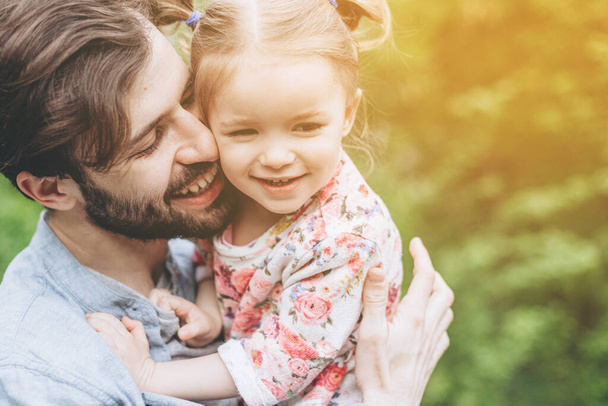 stylish bearded father with his little daughter in his arms against the backdrop of green trees on a walk in the park hugging and kissing a baby - Foto, Imagen