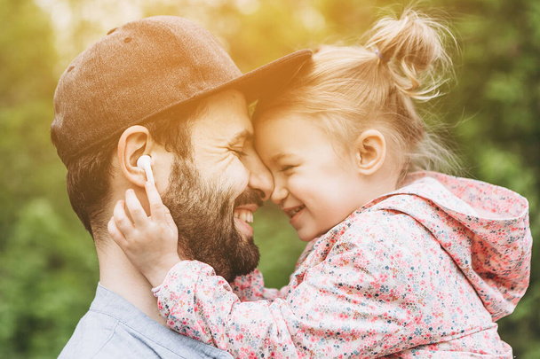 stylish bearded father with his little daughter in his arms against the backdrop of green trees on a walk in the park hugging and kissing a baby - Фото, изображение