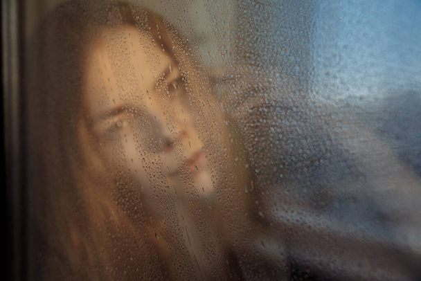 Raindrops trickle down the glass. The window reflects blue sky. The silhouette of girl is visible outside the window. - Фото, зображення