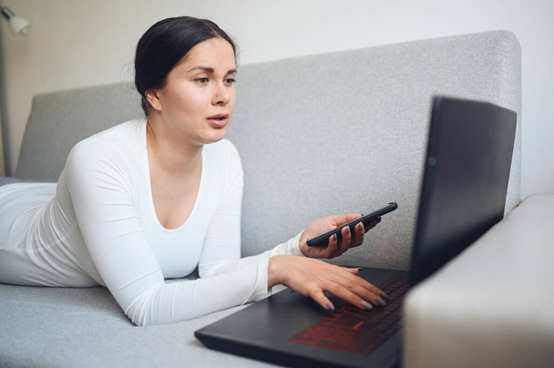 若いヨーロッパのフリーランスの女性は、自宅の隔離中に灰色のソファの上にセルフィーやビデオ通話ノートパソコンや電話を作っています。Covid-19パンデミックコロナウイルス。ホームコンセプトからのオンラインリモートワーク. - 写真・画像