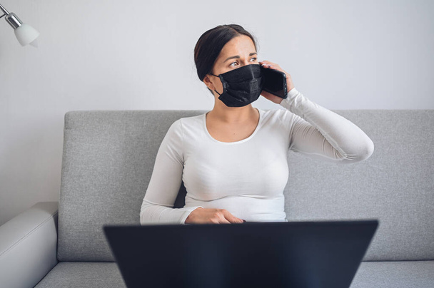 Fiatal szabadúszó nő arcgyógyító maszkban, aki laptopon és telefonon dolgozik szürke kanapén a koronavírus elkülönítés alatt. Covid-19 pandémiás Corona vírus. Online munka otthonról. - Fotó, kép
