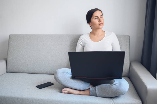 若いヨーロッパのフリーランスの女性は、自宅の隔離中にグレーのソファの上でノートパソコンや電話で働いています。Covid-19パンデミックコロナウイルス。ホームコンセプトからの距離オンライン作業. - 写真・画像