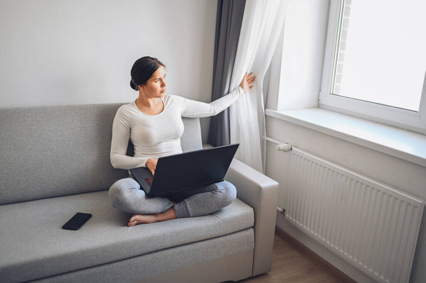 若いヨーロッパのフリーランスの女性は、自宅の隔離中にグレーのソファの上でノートパソコンや電話で働いています。Covid-19パンデミックコロナウイルス。ホームコンセプトからの距離オンライン作業. - 写真・画像