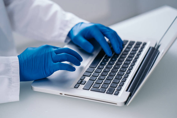 Doctor or scientist in white coat and blue gloves writing on a laptop in his clinic or laboratory office. Biochemistry and biotechnology concept. - Foto, Bild