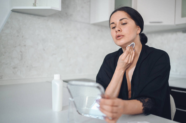 Beautiful brunette woman removing makeup from her face with mirror. Cute beautiful natural beauty girl cleaning face with cotton sponge. Cosmetology and spa, problem skin care, acne treatment concept - 写真・画像