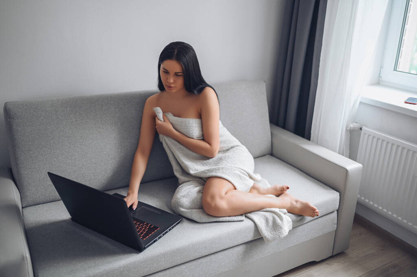 Piękna kobieta korzystająca z laptopa, wykonująca połączenie wideo, pracująca online w domu, pisząca maile, oglądająca telewizję społecznościową lub film, siedząca w wygodnej, szarej kanapie pod ciepłym kocem w domu. Samoizolacja - Zdjęcie, obraz