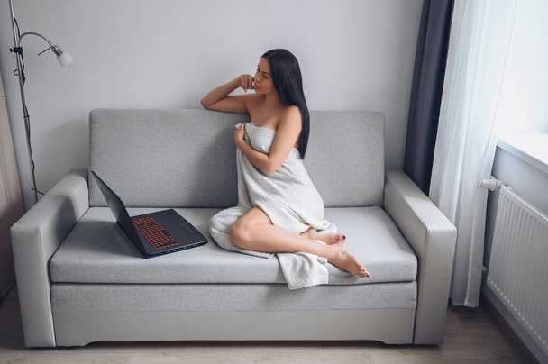Piękna kobieta korzystająca z laptopa, wykonująca połączenie wideo, pracująca online w domu, pisząca maile, oglądająca telewizję społecznościową lub film, siedząca w wygodnej, szarej kanapie pod ciepłym kocem w domu. Samoizolacja - Zdjęcie, obraz