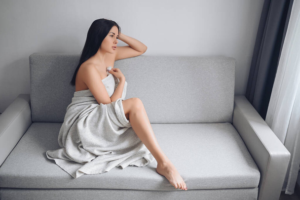 Beautiful bored slender brunette woman sitting in comfortable grey couch under warm blanket at home. Self isolation quarantine during Corona virus pandemic. COVID19 stay home save lives concept - Foto, immagini