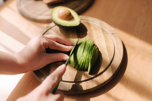 Close up of woman slicing avocado on wooden board at home. Vegetarian food concept - Photo, image