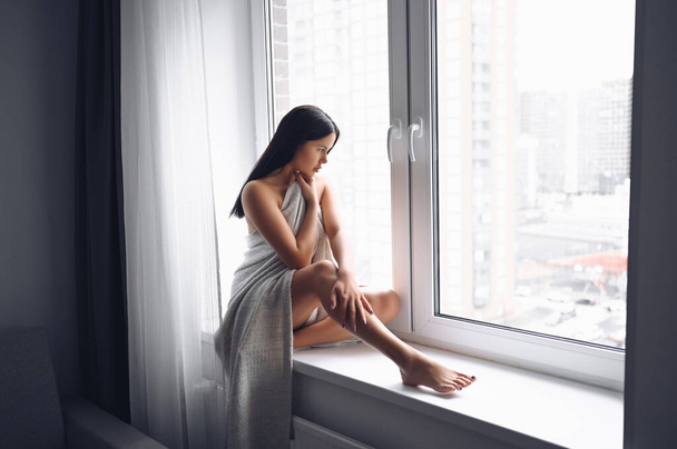 Beautiful bored slender brunette woman sitting next to window windowsill under warm grey blanket at home. Self isolation quarantine during Corona virus pandemic. COVID19 stay home save lives concept - Фото, изображение