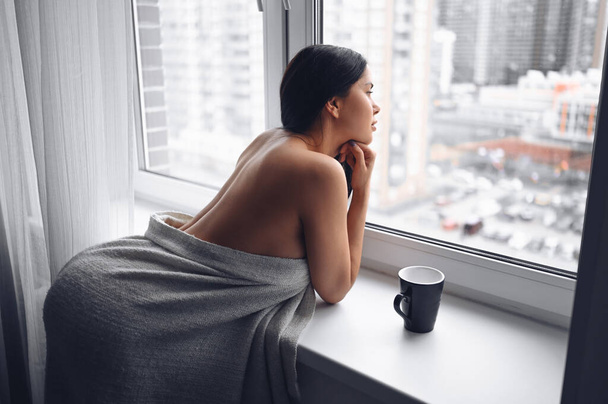 Beautiful bored slender brunette woman sitting next to window windowsill under warm grey blanket at home. Self isolation quarantine during Corona virus pandemic. COVID19 stay home save lives concept - Фото, изображение