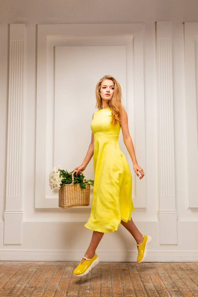 young beautiful  blonde girl in yellow summer dress and yellow shoes with flowers on blue studio background - Foto, afbeelding